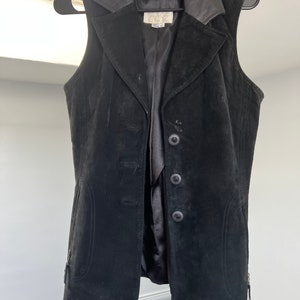 Puede incluir: Chaleco de ante negro con cuello de cuero y cierre de botones. El chaleco tiene dos bolsillos a los lados y una cremallera en cada lado.