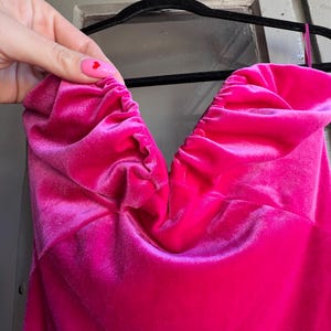 Puede incluir: Un vibrante vestido de terciopelo fucsia con escote coraz&oacute;n. El vestido est&aacute; colgado de una percha negra. La tela est&aacute; fruncida en el escote, creando un efecto texturizado. El vestido es de color liso y parece un vestido formal o de fiesta.