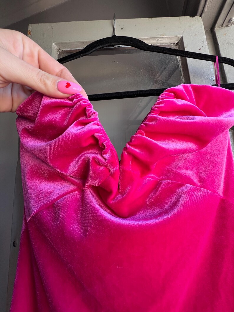 Puede incluir: Un vibrante vestido de terciopelo fucsia con escote coraz&oacute;n. El vestido est&aacute; colgado de una percha negra. La tela est&aacute; fruncida en el escote, creando un efecto texturizado. El vestido es de color liso y parece un vestido formal o de fiesta.