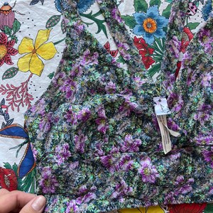May include: A floral bralette in shades of purple, green, and black. The bralette features a deep V-neck and halter straps. The fabric has a textured, lace-like appearance. The garment is displayed on a floral patterned background.