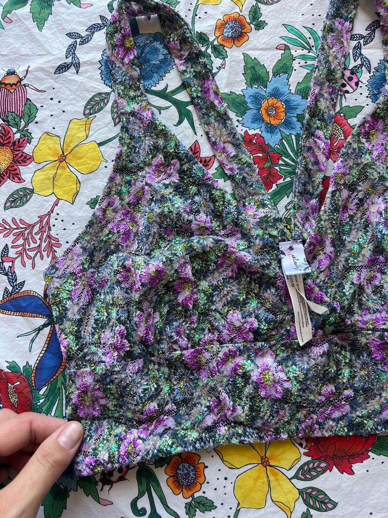 May include: A floral bralette in shades of purple, green, and black. The bralette features a deep V-neck and halter straps. The fabric has a textured, lace-like appearance. The garment is displayed on a floral patterned background.