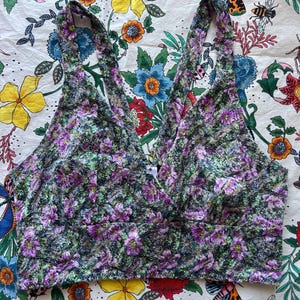 May include: A floral print crop top with a V-neckline. The top features a pattern of purple, green, and white flowers. The straps are wide and the fabric appears to be soft. The background is a floral patterned fabric.