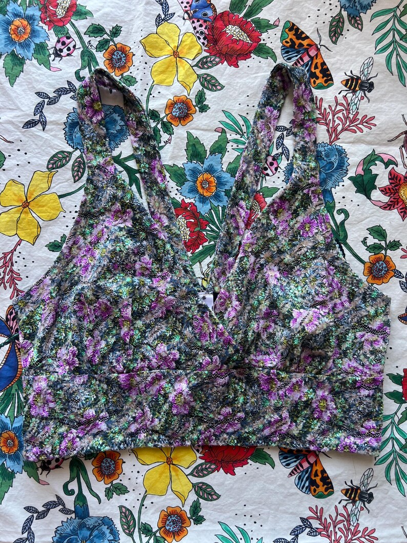 May include: A floral print crop top with a V-neckline. The top features a pattern of purple, green, and white flowers. The straps are wide and the fabric appears to be soft. The background is a floral patterned fabric.