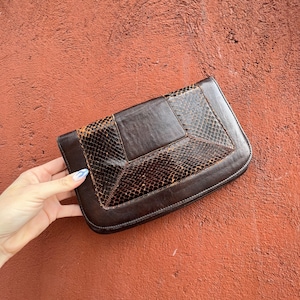 Könnte beinhalten: Eine braune Leder-Clutch mit Schlangenhautmuster. Die Clutch hat einen Klappverschluss und ein geometrisches Design.