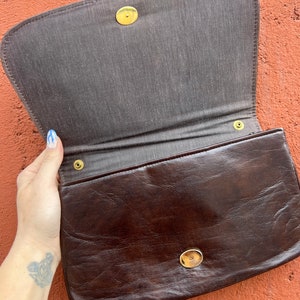 May include: A brown leather clutch bag with a gold clasp and two gold buttons. The bag is open and the inside is lined with a brown fabric.