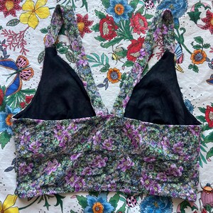 May include: A floral print crop top with black lining and straps. The top features a pattern of purple and green flowers on a dark background. The straps are also adorned with the floral pattern.