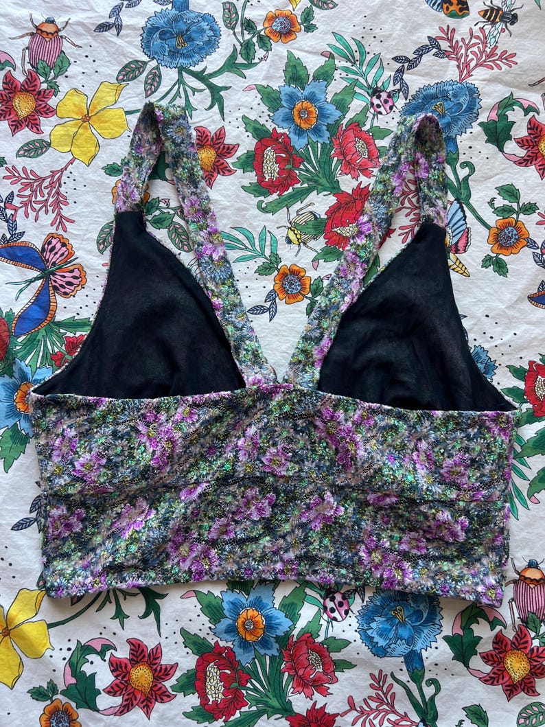 May include: A floral print crop top with black lining and straps. The top features a pattern of purple and green flowers on a dark background. The straps are also adorned with the floral pattern.
