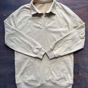 Puede incluir: Una sudadera de color beige de manga larga con cuello de polo y puños de canalé. La sudadera tiene un corte holgado y dos bolsillos de parche en la parte delantera.