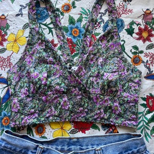 May include: A floral print crop top with a halter neckline and denim shorts. The top features a pattern of purple flowers and green leaves on a black background. The denim shorts are a light blue wash.