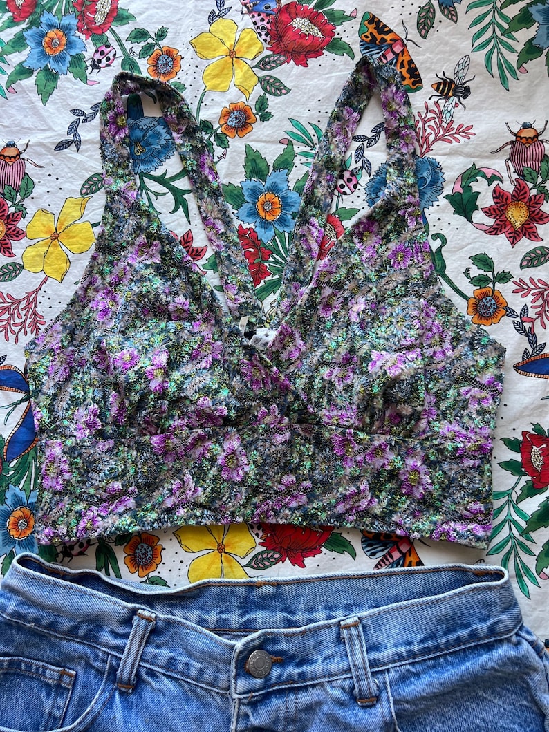 May include: A floral print crop top with a halter neckline and denim shorts. The top features a pattern of purple flowers and green leaves on a black background. The denim shorts are a light blue wash.