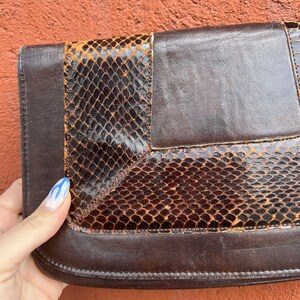 May include: A brown leather clutch with a snakeskin pattern. The clutch has a flap closure and a brown leather strap.