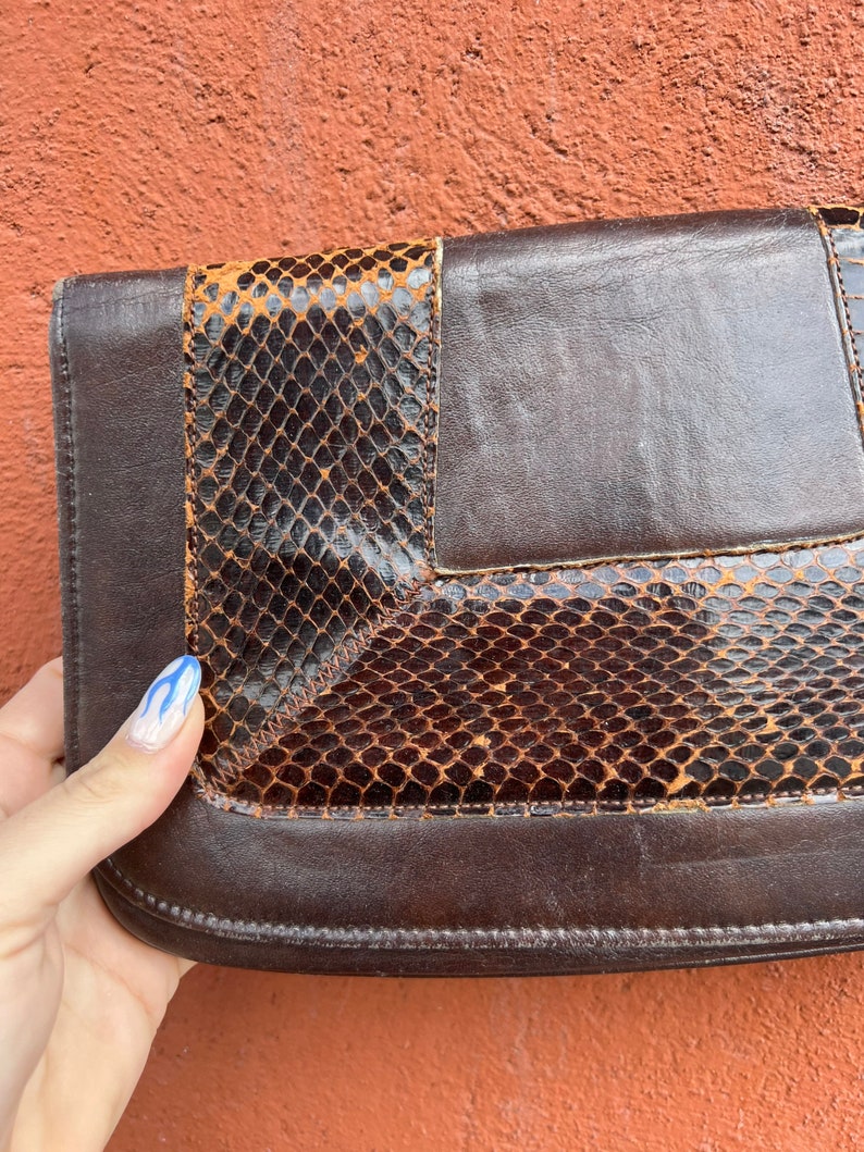 May include: A brown leather clutch with a snakeskin pattern. The clutch has a flap closure and a brown leather strap.
