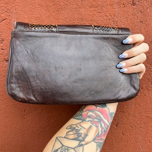 May include: A brown leather clutch bag with a snakeskin pattern on the flap. The bag is held in a hand with blue nail polish.