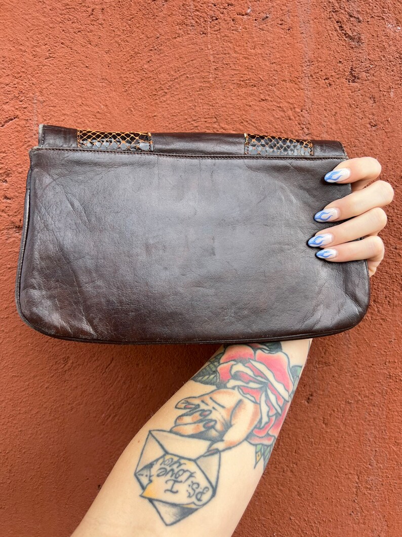 May include: A brown leather clutch bag with a snakeskin pattern on the flap. The bag is held in a hand with blue nail polish.