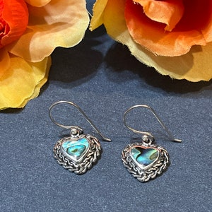 May include: A pair of heart-shaped earrings with abalone shell centers and silver-toned, chain-like borders. The earrings have a hook closure. The background features orange and yellow flowers.