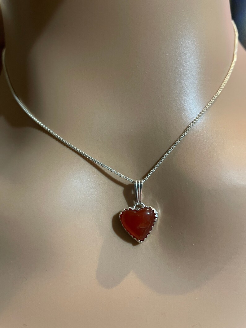 May include: A silver chain necklace with a heart-shaped pendant featuring a red carnelian stone.