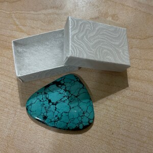May include: A turquoise blue gemstone with a marbled pattern in a white box with a swirling pattern.