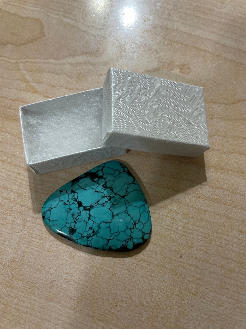 May include: A turquoise blue gemstone with a marbled pattern in a white box with a swirling pattern.