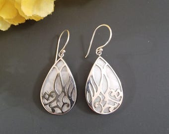 May include: A pair of silver teardrop earrings with a white, pearlescent inlay. The silver frames feature an openwork design. The earrings hang from a simple hook. The background is a dark gray.