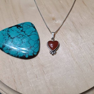 May include: A silver chain necklace with a heart-shaped pendant featuring a red gemstone. The pendant has a silver border with small decorative details.