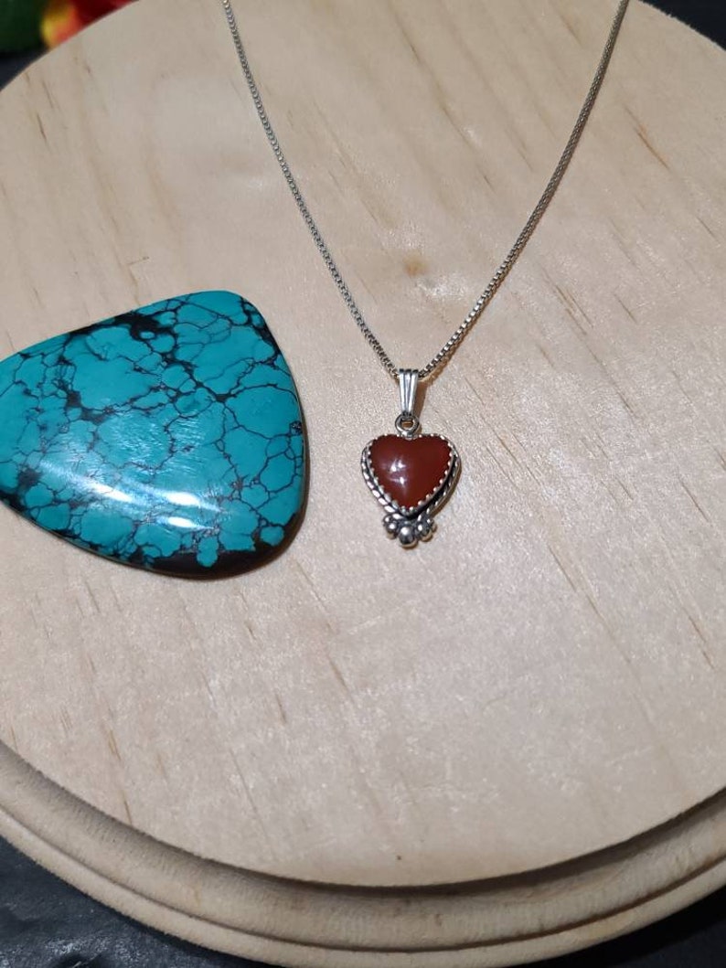 May include: A silver chain necklace with a heart-shaped pendant featuring a red gemstone. The pendant has a silver border with small decorative details.