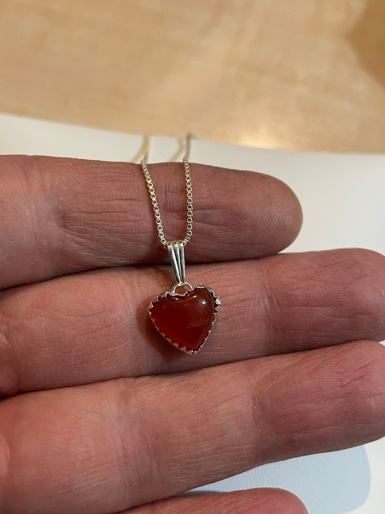 May include: A silver chain necklace with a heart-shaped pendant featuring a red carnelian stone.