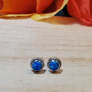 May include: A pair of silver stud earrings with blue opal gemstones. The earrings have a delicate rope-like design around the edges of the gemstones.