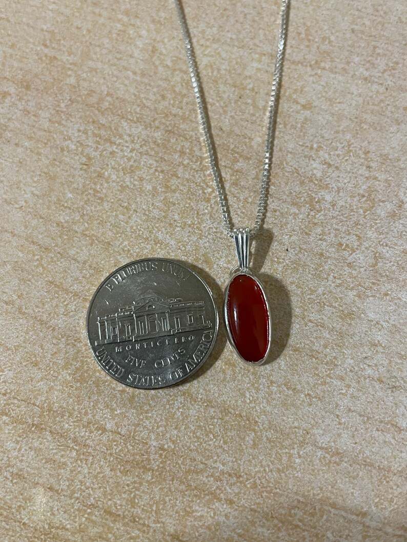 May include: A silver chain necklace with a red carnelian gemstone pendant. The pendant is oval shaped and has a silver setting.