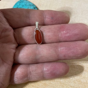 May include: A silver pendant with a red carnelian stone. The pendant is oval shaped and has a simple design.