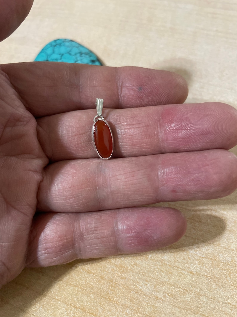 May include: A silver pendant with a red carnelian stone. The pendant is oval shaped and has a simple design.