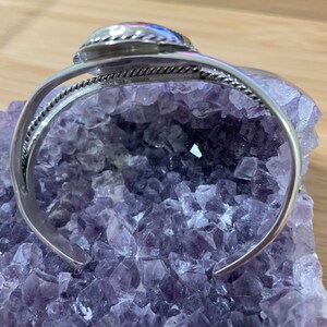 May include: Silver bangle bracelet with a round, silver-toned stone set in the centre. The bracelet is on a bed of purple amethyst crystals.