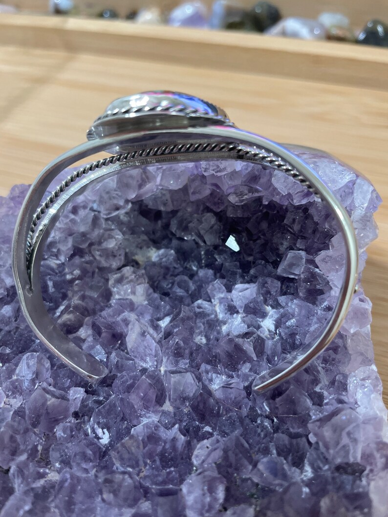 May include: Silver bangle bracelet with a round, silver-toned stone set in the centre. The bracelet is on a bed of purple amethyst crystals.