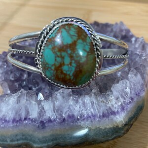 May include: A silver cuff bracelet featuring a turquoise stone set in a rope design. The stone is a mottled green and brown colour.