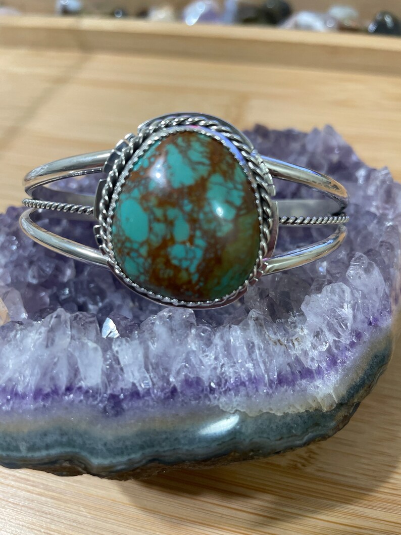 May include: A silver cuff bracelet featuring a turquoise stone set in a rope design. The stone is a mottled green and brown colour.