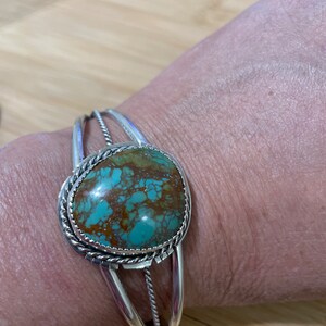 May include: A silver cuff bracelet featuring a turquoise stone set in a rope design. The stone is a mottled green and brown colour.