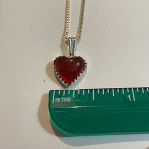 May include: A silver chain necklace with a red heart-shaped pendant. The pendant has a silver border with a scalloped edge.