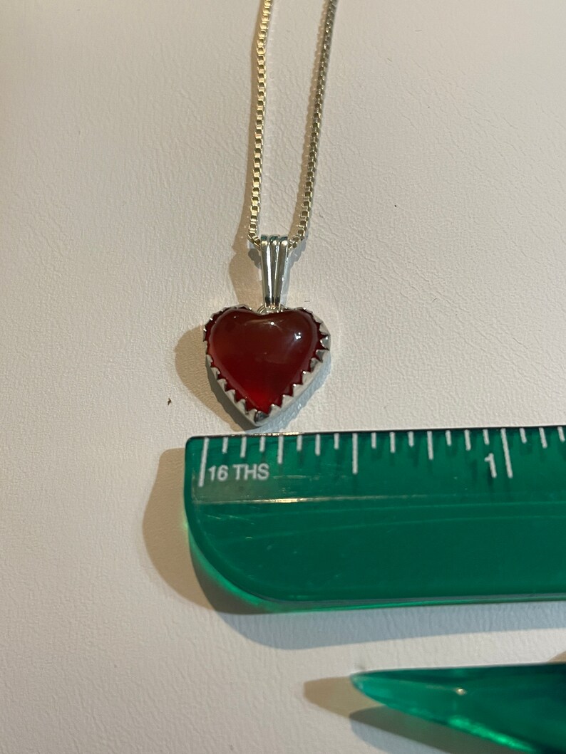 May include: A silver chain necklace with a red heart-shaped pendant. The pendant has a silver border with a scalloped edge.
