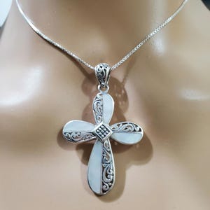 May include: A silver chain necklace with a white mother of pearl cross pendant. The cross has intricate filigree detailing and a small silver square in the center.