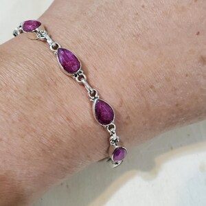 May include: A silver bracelet featuring teardrop-shaped, deep pink gemstones. The bracelet's links are connected by small, decorative silver elements. The gemstones are set in individual silver bezels, creating a delicate and elegant design.