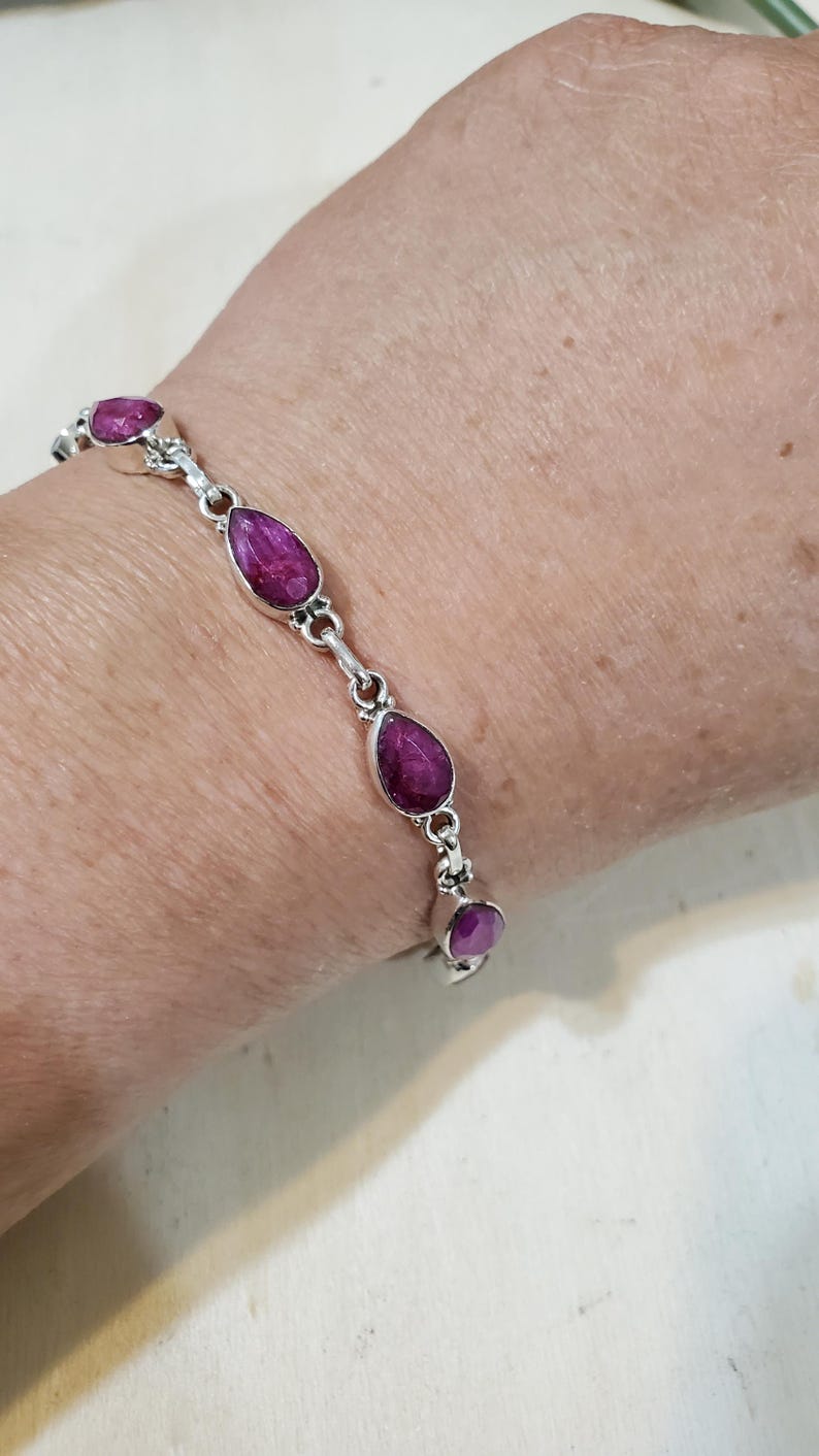 May include: A silver bracelet featuring teardrop-shaped, deep pink gemstones. The bracelet's links are connected by small, decorative silver elements. The gemstones are set in individual silver bezels, creating a delicate and elegant design.