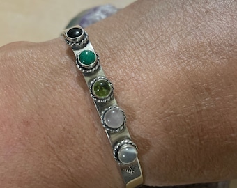 May include: A silver bracelet with a row of colorful gemstones. The stones are set in silver bezels and are surrounded by a twisted silver wire. The bracelet is open and has a clasp.