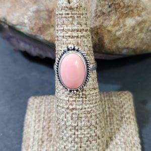 May include: A silver ring with a pink oval gemstone set in a bezel setting. The ring is on a fabric ring display stand.