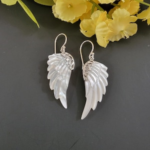 May include: A pair of silver-toned earrings shaped like angel wings. The wings are white with a pearlescent sheen and detailed feathering. The top of each earring features an ornate silver design, and the earrings hang from a curved hook.