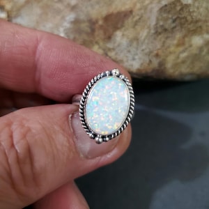 May include: A silver ring with an oval-shaped white opal gemstone set in a silver bezel. The ring has a rope-like design around the bezel and small silver beads around the edge.