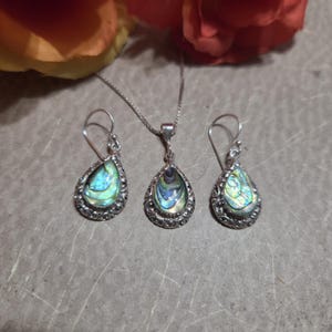 May include: Abalone shell jewelry set featuring a pendant necklace and matching earrings. The teardrop-shaped pendants and earrings have a silver-colored filigree border. The abalone shells display iridescent blues, greens, and purples.