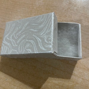 May include: A small, white, rectangular box with a swirl pattern on the lid. The box is open and the inside is lined with a soft, white material.