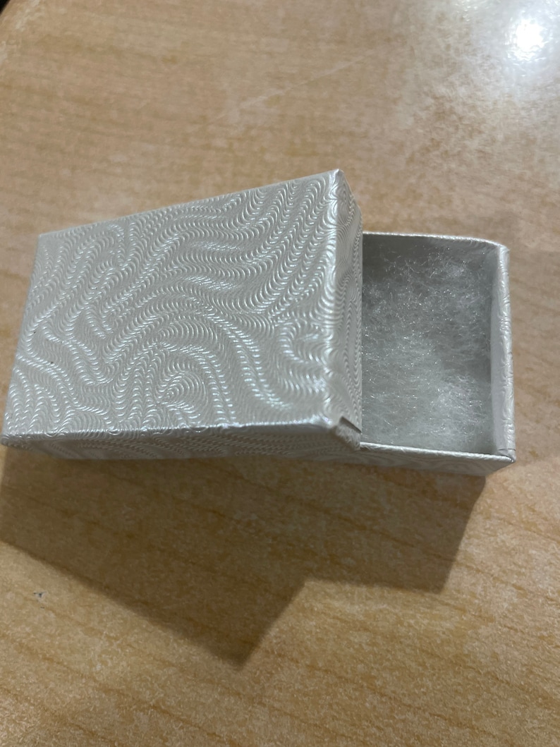 May include: A small, white, rectangular box with a swirl pattern on the lid. The box is open and the inside is lined with a soft, white material.