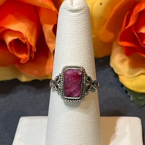 May include: A silver ring with a pink rectangular gemstone set in a decorative bezel. The ring is on a white ring display stand.