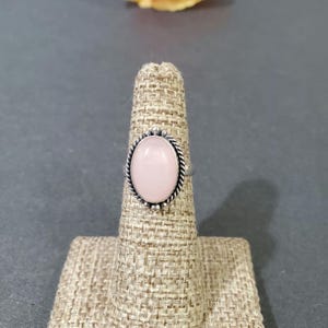 May include: A silver ring with a pink oval gemstone set in a decorative bezel. The ring is displayed on a fabric ring holder.