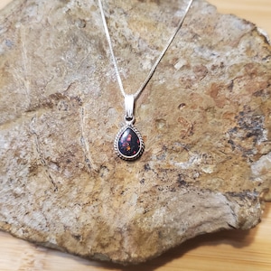 May include: A silver necklace with a teardrop-shaped pendant. The pendant features a dark stone with red and black flecks, surrounded by a silver rope design. The necklace is displayed on a textured, brown stone.